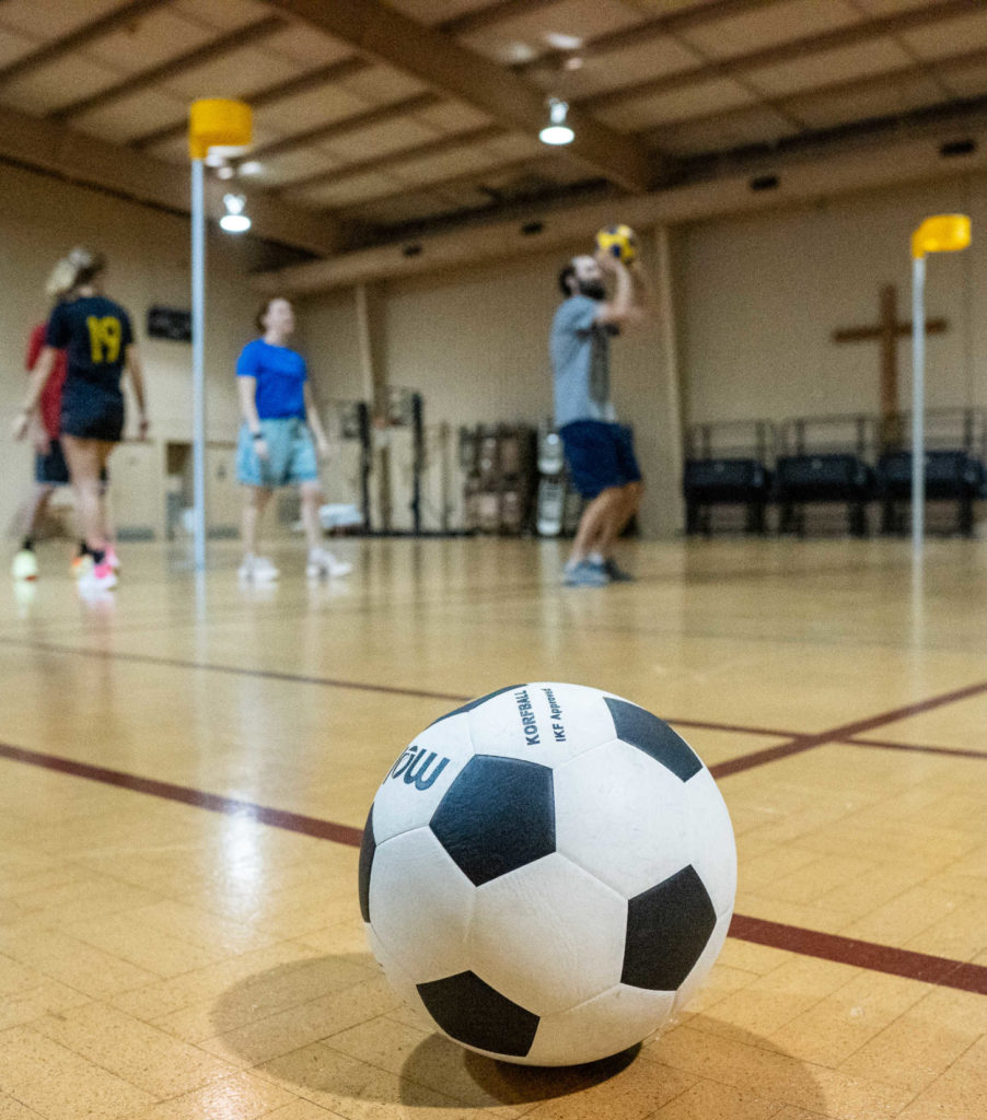 Members of the US National Team practice korfball with one another for the first time. One member is beginning to shoot the korfball while three others are watching. There is a korfball in the foreground at the center of the picture.